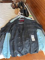 Black and gray patterned short-sleeve shirt hanging on white hanger, with light blue shirt beneath it, placed on bed with three pairs of men's shoes lined up behind.