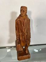 Front and slightly angled view of the wooden carved man figurine wearing a long coat and hat, holding a bag