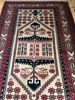 Full view of rectangular Baluchi rug with central geometric medallion and floral motifs, and multiple patterned borders in colors black, pink, beige, and dark red. Fringed edges on shorter sides visible.