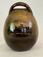 Front view of vase showing the handle and a scenic painting of buildings in dark tones on glossy brown ceramic.