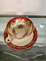 Aynsley tea cup and saucer displayed together on a glass surface; tea cup has floral interior and red border with gold filigree.