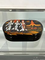 Closed oval jewelry box with black lacquer finish and hand-painted traditional scene on lid featuring three women and pagoda.