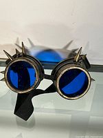 Front view of goggles showing round blue lenses with bronze-colored spiked frames and black strap.