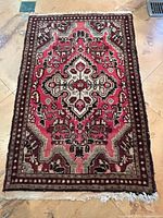 Full view of rectangular wool rug showing medallion design and patterned borders in red, ivory, black and green tones. Fringe visible at bottom edge.