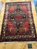 Full view of the rectangular Mazlagan wool rug showing the overall design with a central dark medallion and contrasting cream border on a red background.