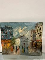 Front view of the signed oil on canvas painting showing urban street scene with people walking along wet street and buildings under cloudy sky.