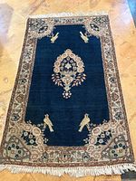 Full top-down view of rectangular blue wool rug with central medallion, bird motifs, floral border and fringed ends on tiled floor