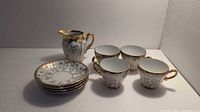 Photo of four tea cups with gold floral details, matching saucers stacked, and creamer jug on a white surface