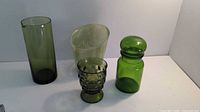Photo showing four different green glass pieces on a white surface: two vases, one patterned glass cup, and one jar with stopper.