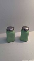 Pair of green glass salt and pepper shakers with silver metal tops on white surface against plain background.