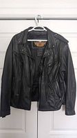 Front view of black leather Harley Davidson motorcycle jacket hanging on a white hanger.
