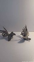 Two heavy metal rooster figurines viewed from above showing wing and feather details
