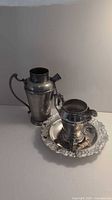 Tall silver tone pitcher, creamer on ornate round tray on white background