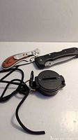 Photo showing both pocket cutters and the engineer lensatic compass with lanyard on a white surface.