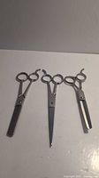 Three professional hair cutting scissors laid out on a white surface: two thinning scissors and one regular cutting scissor, silver metallic finish