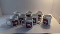 Eight white ceramic coffee mugs arranged in two rows, each featuring a red Illy logo on the side.