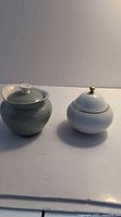 Two sugar bowls shown on flat white surface, one grey rounded with lid, one white textured with gold knob lid