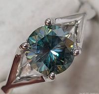 Close-up front view of silver ring with round blue-green moissanite gemstone held by four prongs.