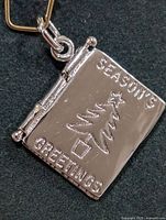 Front cover of the polished sterling silver book pendant, engraved with a Christmas tree and the phrase "SEASONS GREETINGS."
