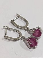 Pair of sterling silver earrings with oval red ruby gemstones and small clear stones set on the hoops, showing front and side views.