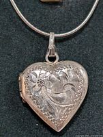 Close-up image of the silver heart-shaped locket necklace showing detailed engraved floral and leaf patterns on the front side.