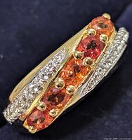 Close-up angled view of the curved ring showing five round orange sapphires set in gold-toned prongs with clear stones on either side in a silver band.