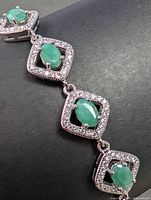 Close-up photo showing the oval green emerald stones each framed by a square of small clear cubic zirconia stones set in silver metal.