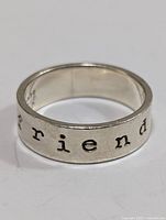 Close-up image of silver band ring showing the word 'friend' engraved around the band in typewriter style font, size 7.