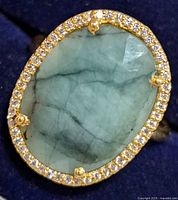 Close-up of oval green emerald set in gold-tone bezel surrounded by small clear stones.