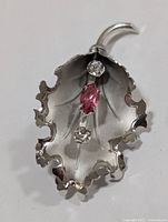 Close-up image of a silver leaf-shaped brooch with a marquise-cut pink stone and two clear cubic zirconia stones on the leaf veins. The brooch has a curled stem.