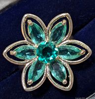Close-up view of the flower-shaped silver ring with six marquise green gemstones and a round central green gemstone, displayed on a dark blue background.