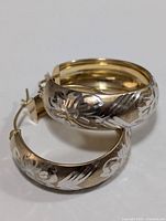 Close-up image of two silver hoop earrings featuring floral and leaf pattern engraving on the exterior and a yellow-gold polished interior surface.