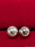Close-up photo of two silver ball stud earrings, showing size difference and shiny surfaces on red velvet background.