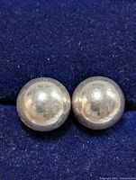 Close-up image of two silver dome-shaped earrings placed side by side on a dark blue fabric background. The surface is slightly reflective with signs of wear.
