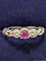 Front close-up of silver ring with central ruby and two small clear side stones, showing the intertwined band design.