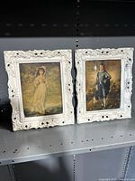 Two antique pictures with ornate white carved frames displayed side by side on a shelf.