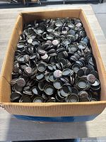 Wide top view of a large cardboard box filled with many unused Coca-Cola bottle caps.