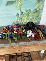 Wide shot showing a variety of action figures from multiple franchises including Marvel, Star Wars, Transformers, and Teenage Mutant Ninja Turtles arranged on a wooden table.