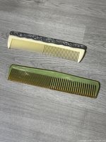 Photo showing both antique celluloid combs placed side by side on a grey wood floor. The top comb is cream-colored with a silver decorative metal strip and fine teeth. The bottom comb is green marbled with wide teeth.