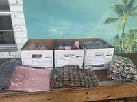 Three bankers boxes containing various pieces of folded clothing and stacks of folded apparel with different colors and patterns shown on a wooden table.