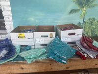 Three bankers boxes filled with folded new clothing items with additional garments laid out in front on a wooden table. Clothing colors include teal, red, blue, maroon, grey, and mint green.