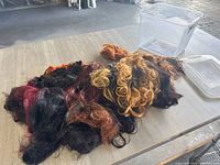 Wigs and hair extensions laid out on a surface next to clear plastic storage containers, showing mixed colors including black, red, blonde, and brown with curly and wavy texture.