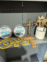 Shelf display showing two round metal wall plates with text and turquoise truck image, yellow metal scroll piece, and two wooden crosses plus ceramic vase with floral arrangement