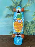 Full view of the skateboard showing the deck with pineapple graphic, trucks, and orange wheels.