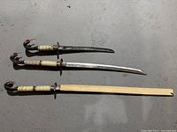 Full view of three cobra swords laid out side by side on concrete floor showing blades, handles, and scabbards.