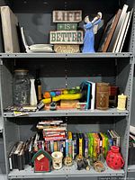 Wide view of shelving unit with mixed items: DVDs, VHS tapes, home decor signs, glass jar, wooden pepper mill, assorted knickknacks and dishes