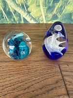 Two blown glass paperweights on wooden surface with palm leaves background. Blue egg-shaped paperweight with white internal swirl on the right; clear spherical paperweight with blue internal detail on left.