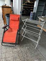 Photo showing orange outdoor lounger next to white folding clothes drying rack on paved area indoors.