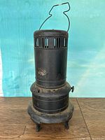 Black cylindrical metal heater stove with wire handle, vent openings near the top, rounded base on three feet, latch on side, shown from front.