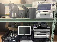 Photo showing shelf with coffee maker, printers, speakers, IBM Trackpoint II keyboard box, and some stereo receivers on the right side. Plastic bin with cables below shelf along with a flat screen monitor and stacked electronic components.